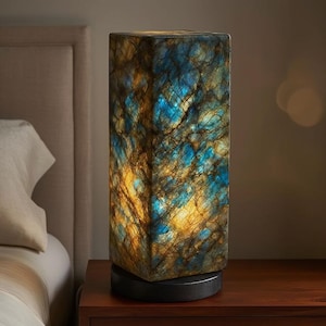 May include: A rectangular table lamp with a distinctive, textured surface. The lamp displays a mix of blue, gold, and brown tones, producing a striking visual effect. It rests on a dark, circular base and is positioned on a wooden bedside table.
