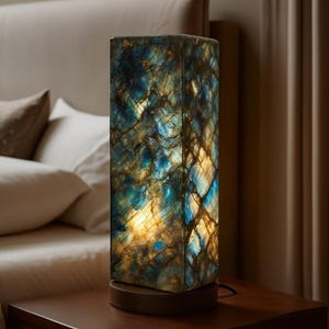 May include: A rectangular table lamp with a unique, patterned design. The lamp features a base and a tall, rectangular shade with a blue, gold, and brown pattern. The lamp is on a wooden nightstand next to a bed.