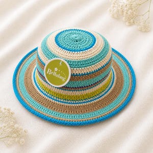 Peut inclure: Un chapeau de soleil en crochet à larges rayures. Le chapeau présente des rayures alternées bleues, turquoise, beiges, marron et vertes. Une étiquette ronde avec le texte "Bellabug" est attachée.