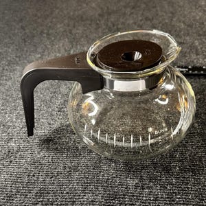 May include: A white ceramic mug with a black handle and the text "But First, Coffee" in a black, handwritten-style font. The mug is on a white surface.