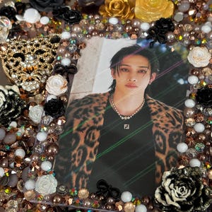 May include: A decorative photo frame with a picture of a person wearing a leopard print jacket. The frame is embellished with black, gold, and white roses, pearls, and rhinestones. A gold-colored leopard head is on the left side of the frame.