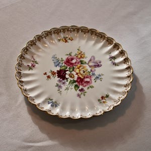 May include: A white plate with a scalloped edge, decorated with a floral bouquet. The bouquet includes pink, red, yellow, and purple flowers. The plate has a gold trim around the edge.