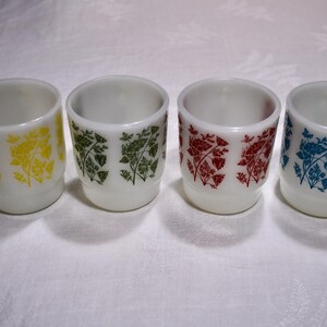 May include: Four white milk glass tumblers arranged in a row. Each tumbler features a repeating floral pattern printed in a different colour: yellow, green, red, and blue. The cups have a cylindrical shape with a slightly wider rim and a base.