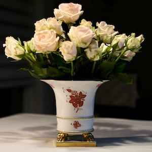 Puede incluir: Un jarrón de cerámica blanco con un diseño floral contiene un ramo de rosas de color rosa claro. El jarrón tiene una parte superior acampanada, una banda decorativa y descansa sobre una base cuadrada dorada con patas de león. Las rosas tienen hojas verdes.