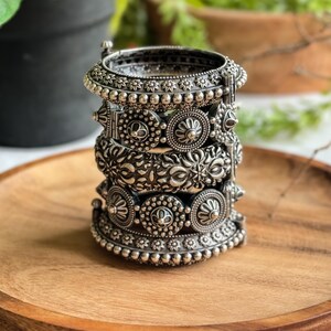 Könnte beinhalten: Ein Stapel aus vier silberfarbenen Metallarmreifen mit komplexen geprägten Blumen- und geometrischen Mustern. Jeder Armreif zeigt ein eigenes strukturiertes Design mit kleinen Perlenrändern und einer metallischen Oberfläche.