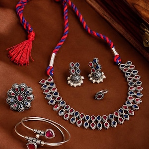 Könnte beinhalten: Ein Schmuckset bestehend aus einer Halskette mit blau-roter Kordel, einem Paar Jhumka-Ohrringen, einem blumenförmigen Ring, einem kleineren Ring und einem Armreif, gefertigt aus oxidiertem silberfarbenem Metall mit roten und blauen Steinen.