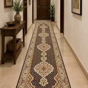 May include: A long, narrow runner rug featuring a dark brown background with intricate cream, tan, and rust-colored floral and geometric patterns. The rug has a symmetrical design with repeating central medallions and a detailed border along the edges.