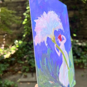 Puede incluir: Pintura original sobre lienzo que representa una figura que se extiende hacia una gran flor blanca y rosa sobre un fondo azul. La obra de arte presenta a una mujer con un vestido blanco y cabello rojo, rodeada de follaje verde. La pintura se sostiene en &aacute;ngulo.