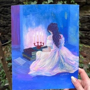 Op de afbeelding: Een prent van een vrouw in een witte jurk, zittend bij een kandelaar met brandende kaarsen en boeken. Het kunstwerk heeft een blauw en paars kleurenschema, met de rug van de vrouw naar de kijker. De prent wordt door een hand vastgehouden.