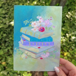 May include: A stack of vintage books with a bouquet of pink and white flowers on top. The books are painted in shades of blue, purple, and white, with gold accents. The flowers are painted in a realistic style, with delicate petals and a vibrant red center.
