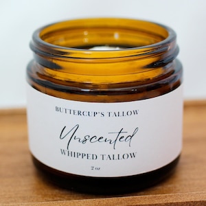 Op de afbeelding: Een kleine, amberkleurige glazen pot met een wit etiket. Het etiket luidt "Buttercup's Tallow Unscented Whipped Tallow 2 oz." De pot heeft een donkerbruine deksel en staat op een houten oppervlak.