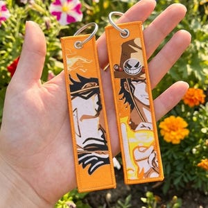 Könnte beinhalten: Zwei orangefarbene, rechteckige Schlüsselanhänger mit silbernen Schlüsselringen. Jeder Schlüsselanhänger zeigt eine Charakterillustration in Braun-, Weiß- und Schwarztönen. Der Hintergrund ist leuchtend orange.