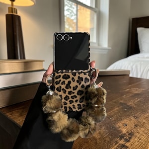 Könnte beinhalten: Eine Smartphone-Hülle mit Leopardenmuster. Die Hülle hat einen schwarzen Rahmen und eine Klappe mit passendem Leopardenmuster. Sie verfügt über braune Fellbommel, die mit einem Perlenakzent an der Hülle befestigt sind.