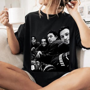 May include: A black short-sleeved t-shirt featuring a monochromatic graphic print of four individuals in athletic gear. The design shows the group in a close-up composition, with one person wearing a glove labeled with the text WARRIOR.