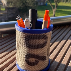 Könnte beinhalten: Ein zylindrischer Stifthalter mit blauem Rand und Boden, umwickelt mit beigem Stoff mit dunklem Muster. Der Halter enthält mehrere Stifte und Marker in Orange, Weiß und Schwarz. Der Halter steht auf einer Holzoberfläche.