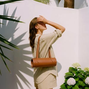 May include: A tan-brown cylindrical leather handbag with two long straps and silver rivets. The bag is carried by a person wearing a beige linen shirt and shorts. The background features a white wall and green foliage.