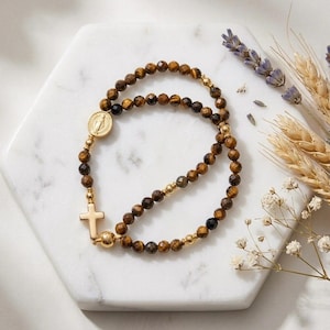 May include: A beaded bracelet featuring faceted brown tiger eye stones, small gold-toned metal spacer beads, a circular gold-toned medal depicting a figure, and a small gold-toned cross charm. The bracelet is arranged in a circular loop.