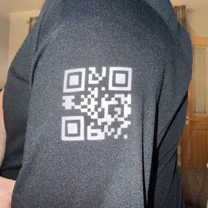 May include: Black short-sleeved shirt featuring a white QR code on the sleeve. The fabric has a textured appearance. The shirt is likely designed for sports or physical activity.