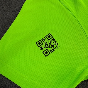 May include: Close-up of a neon green athletic shirt. The fabric has a textured appearance. A black QR code is printed on the shirt's lower left side. The shirt's seams are visible, indicating its construction.