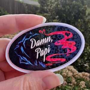 May include: Oval sticker with the text "Damn, Papi" in white script. The sticker features a blue and black design on the left and a red number "3" on the right, with a heart-shaped design. The sticker has a white border.
