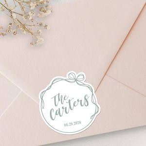 May include: A pale pink envelope sealed with a circular sticker featuring a sage green line-art ribbon border, the text "The Carters" in a script font, and the date "20.06.2026" printed in a simple sans-serif typeface at the bottom.