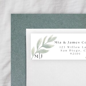 Puede incluir: Una etiqueta de dirección rectangular con una ilustración de hojas verdes en acuarela, las iniciales M|J y el texto impreso Mia & James Collins, 123 Willow Lane, San Diego, CA 92101, colocada sobre un sobre blanco y una cartulina verde salvia.