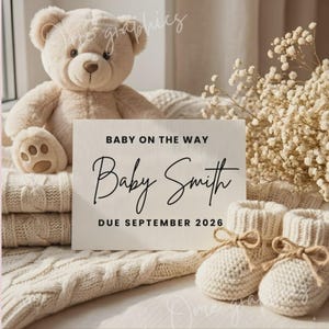 May include: A beige plush teddy bear sits behind a white rectangular sign with black text that reads BABY ON THE WAY, Baby Smith, DUE SEPTEMBER 2026. A pair of cream-colored knit baby booties with brown twine bows sit next to a bouquet of dried flowers.