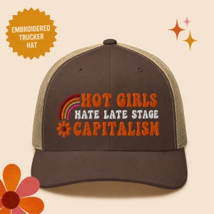 May include: Brown trucker hat with a tan mesh back. The hat features the embroidered text "HOT GIRLS HATE LATE STAGE CAPITALISM" with a rainbow and flower design. The hat also has the text "EMBROIDERED TRUCKER HAT".