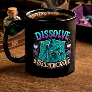May include: A black ceramic mug filled with coffee, featuring the text "DISSOLVE GENDER ROLES" in a colorful, fantasy-themed design. The design includes a skeleton, dice, and a translucent cube. Steam rises from the coffee, suggesting it is hot.