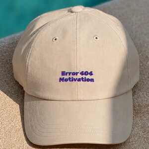 Peut inclure: Une casquette de baseball beige avec le texte brodé Error 404 Motivation en lettres violettes. La casquette présente une calotte structurée avec des œillets de ventilation et une visière incurvée, offrant un design simple et épuré.