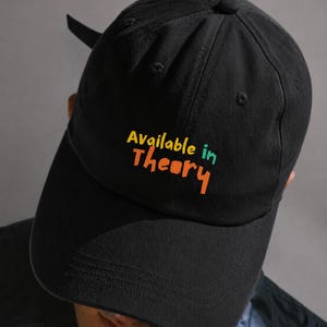 Op de afbeelding: Een zwarte baseballpet met de tekst Available in Theory geborduurd op de voorzijde. Het woord Available is geel, het woord in is blauwgroen en het woord Theory is oranje. De pet heeft een gebogen klep en een gestructureerde kroon.