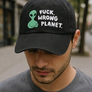 May include: A black baseball cap featuring a graphic of a light green alien with crossed arms and the white text FUCK. WRONG PLANET. The cap has a curved brim, a button on top, and stitched eyelets for ventilation on the crown panels.