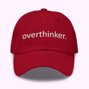 Puede incluir: Gorra de béisbol roja con la palabra "overthinker." en blanco, centrada en la parte delantera. La gorra tiene una visera curva y un botón en la parte superior. El sombrero es de color liso y parece estar hecho de una tela suave.