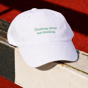 Può includere: Cappellino da baseball bianco con la scritta verde "Thinking about not thinking". Il cappellino è appoggiato su una superficie di legno usurata, con uno sfondo rosso. Il cappellino ha una visiera curva e un bottone sulla parte superiore.