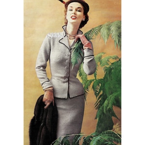 May include: A woman wearing a gray knitted suit with a black fur stole. She is standing in front of a leafy plant and wearing a red hat with a feather.