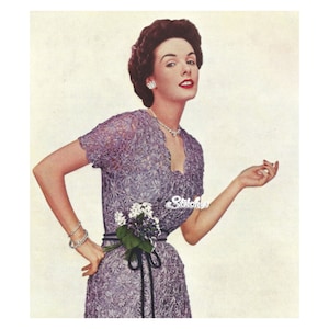 May include: A woman wearing a purple crochet dress with a black sash and a bouquet of white flowers. The dress has a lace pattern and a V-neckline. The woman is smiling and looking at the camera.