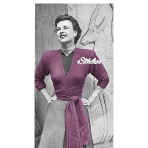 May include: A woman wearing a pink wrap sweater with a matching fringed scarf tied around her waist. The sweater has a textured knit pattern.
