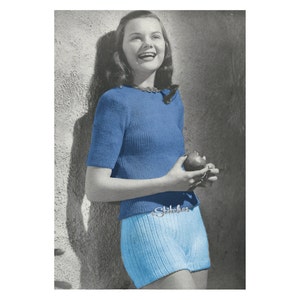 May include: A woman wearing a blue knitted sweater and matching blue knitted shorts. She is smiling and holding a red apple.