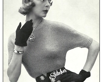 May include: A woman wearing a gray knitted sweater with a black belt that says "e Stitches". She is wearing black gloves and has her hair styled in a short, curled bob.