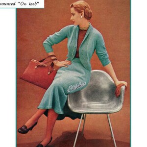 May include: A woman wearing a teal blue knitted cardigan and skirt set, sitting on a silver chair. She is holding a red handbag and has a pearl earring in her right ear. The text "ANTIBES" is printed in black at the top of the image, with the text "pronounced "On teeb"" printed below it.
