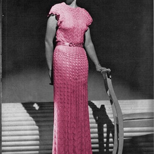 Puede incluir: Una mujer con un vestido de crochet rosa con un dobladillo festoneado. El vestido tiene un corpiño ajustado y una falda completa. La mujer está de pie junto a una silla y mira hacia un lado.