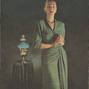 Può includere: Una donna in un vestito verde con una gonna drappeggiata sta in piedi davanti a uno sfondo scuro. Indossa gioielli d'oro e tiene le mani giunte davanti a sé.