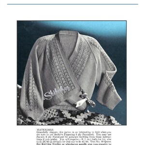 May include: A gray knitted cardigan with a lace pattern. The cardigan has a wrap style and a tie at the waist. The cardigan is laying on a white crocheted fabric.