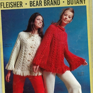 Puede incluir: Dos mujeres modelando suéteres de punto. La mujer de la izquierda lleva un suéter blanco de punto de cable con flecos. La mujer de la derecha lleva un suéter rojo de punto de cable con flecos. Ambos suéteres tienen botones.