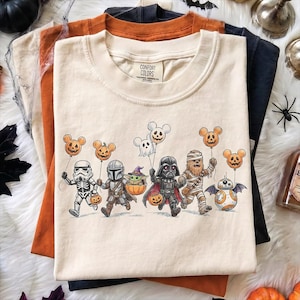 May include: A cream-colored t-shirt with a graphic print featuring Star Wars characters dressed for Halloween, including a Stormtrooper, Mandalorian, Grogu, Darth Vader, Chewbacca, and BB-8, holding pumpkin and ghost balloons with mouse ears.