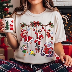May include: A cream-colored t-shirt featuring a graphic print of various animated characters dressed in Christmas attire, including a mermaid, a prince, a crab, a seagull, a fish, and a sea witch, all hanging from a decorated tree branch.