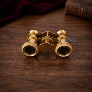 以下が含まれることがあります： 真鍮製のアンティークオペラグラス。双眼鏡は、中央のフォーカスホイールとアイカップを備えたクラシックなデザインです。真鍮は研磨仕上げです。背景には木製の表面と濃い赤色のベルベットが含まれています。