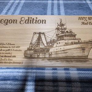 Könnte beinhalten: Holzbrett mit der Aufschrift "Oregon Edition" und einer detaillierten Gravur eines Fischerbootes. Das Brett besteht aus 100 % Western Red Cedar und misst etwa 30 cm. Gebrauchsanweisungen sind ebenfalls enthalten.
