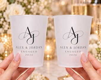 May include: Two frosted plastic cups featuring a black printed monogram of the letters AJ with a decorative flourish. Below the monogram, the text reads ALEX & JORDAN, ENGAGED, and 2026 in a clean, black serif font centered on each cup.