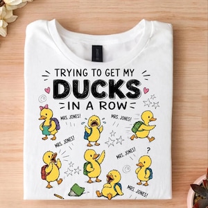 Könnte beinhalten: Ein weißes T-Shirt mit einer Grafik von sechs gelben Entenküken mit Rucksäcken und dem Text "TRYING TO GET MY DUCKS IN A ROW" sowie "MRS. JONES!", der um die Illustrationen verteilt ist. Das Design enthält kleine Sterne, Wirbel und einen Bleistift.